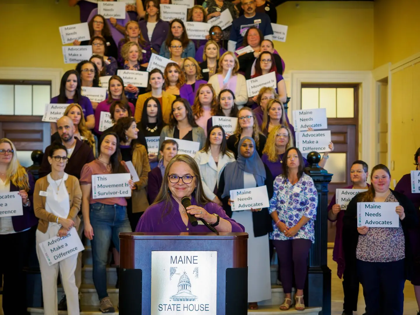 Francine Garland Stark speaking at a Maine State House podium with advocates holding signs behind her