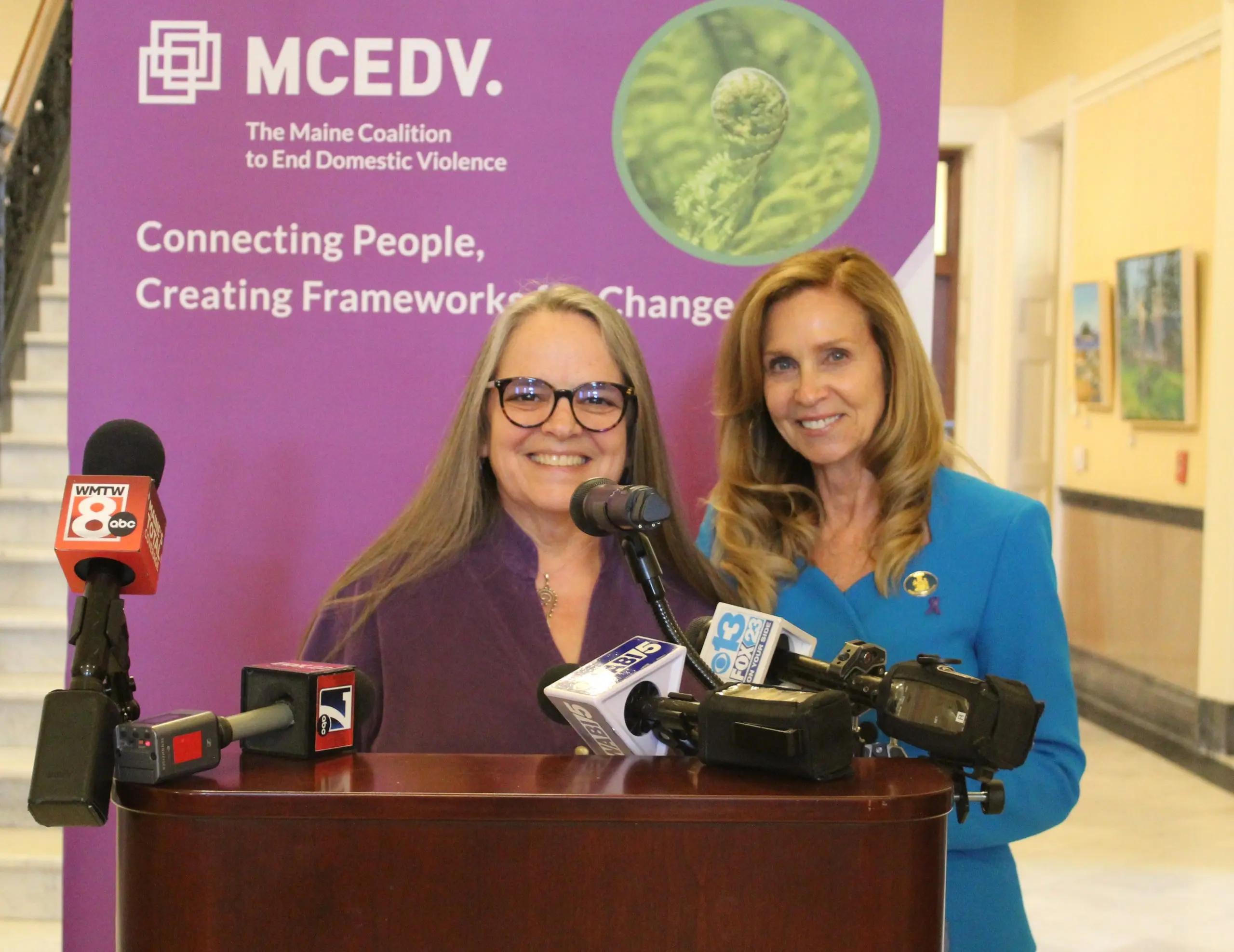 Two women smiling at a podium crowded with news microphones, in front of an MCEDV banner